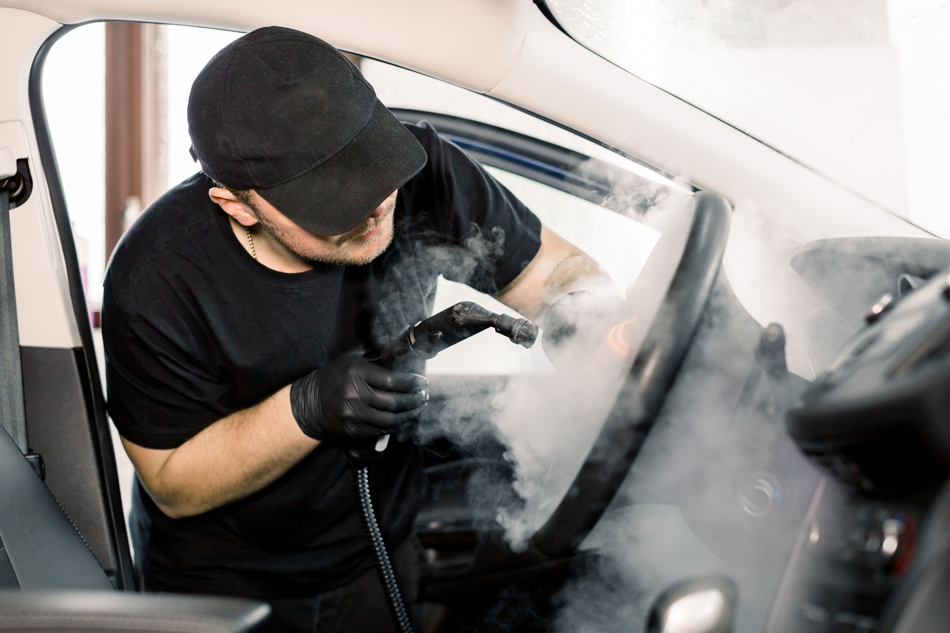 Car detailing, steam cleaning concept. Handsome man in black t-shirt and cap, worker of car wash center, cleaning car interior with hot steam cleaner. Car detailing concept